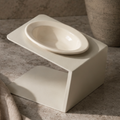 White ceramic dish on a matching stand with a textured vase in the background.